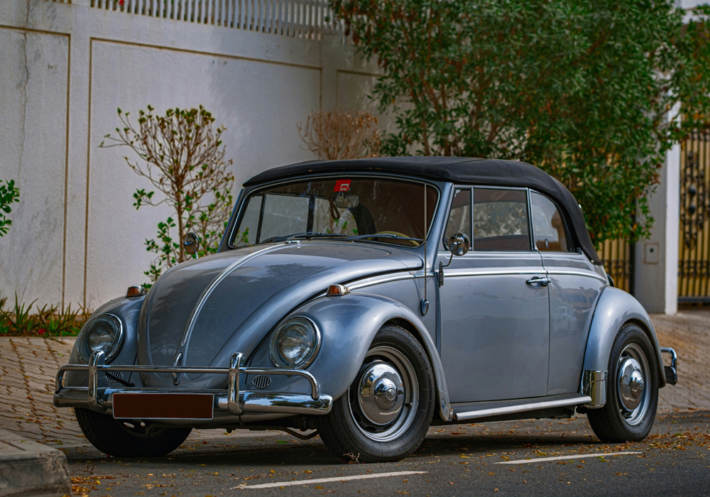 Silver blue Volkswagen Beetle convertible parked beside a white wall with trees and greenery